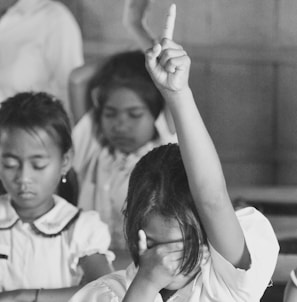 A classroom setting with three children sitting. One child has their hand raised with one finger pointing up, while the other hand covers their face. The other two children have their eyes closed, appearing focused or contemplative.