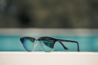 A pair of stylish sunglasses with dark frames and reflective blue lenses is placed on a white surface. The background is blurry, primarily showing a turquoise-colored pool or body of water.