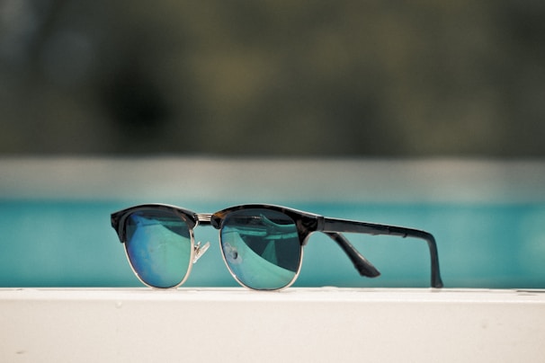 A pair of stylish sunglasses with dark frames and reflective blue lenses is placed on a white surface. The background is blurry, primarily showing a turquoise-colored pool or body of water.
