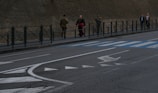 A city street scene with a group of people walking on the sidewalk beside a stone building. Two individuals wear military uniforms, and one person in clerical attire with a red sash walks near them. Other pedestrians follow, and the road has painted markings including arrows and a pedestrian crossing.