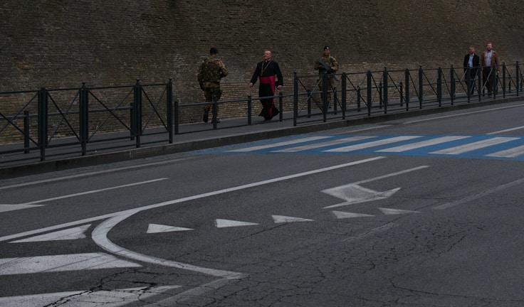 A city street scene with a group of people walking on the sidewalk beside a stone building. Two individuals wear military uniforms, and one person in clerical attire with a red sash walks near them. Other pedestrians follow, and the road has painted markings including arrows and a pedestrian crossing.
