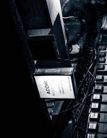 A monochrome image shows a collection of rectangular signs and a network of intertwined cables. The most prominent sign is illuminated with the text 'ACOHI Crafted Objects Concept Store.' The environment conveys a sense of urban chaos with a focus on the industrial and mechanical elements of the scene.
