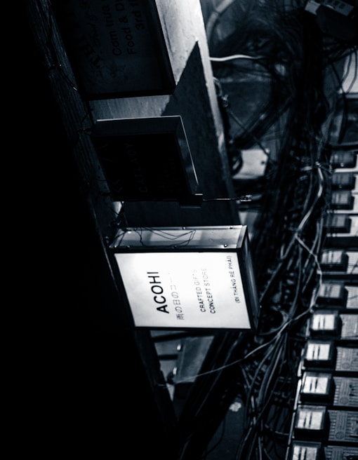 A monochrome image shows a collection of rectangular signs and a network of intertwined cables. The most prominent sign is illuminated with the text 'ACOHI Crafted Objects Concept Store.' The environment conveys a sense of urban chaos with a focus on the industrial and mechanical elements of the scene.