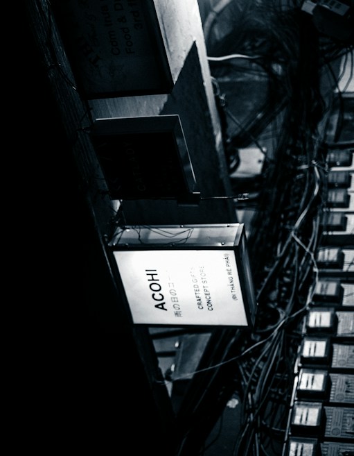 A monochrome image shows a collection of rectangular signs and a network of intertwined cables. The most prominent sign is illuminated with the text 'ACOHI Crafted Objects Concept Store.' The environment conveys a sense of urban chaos with a focus on the industrial and mechanical elements of the scene.