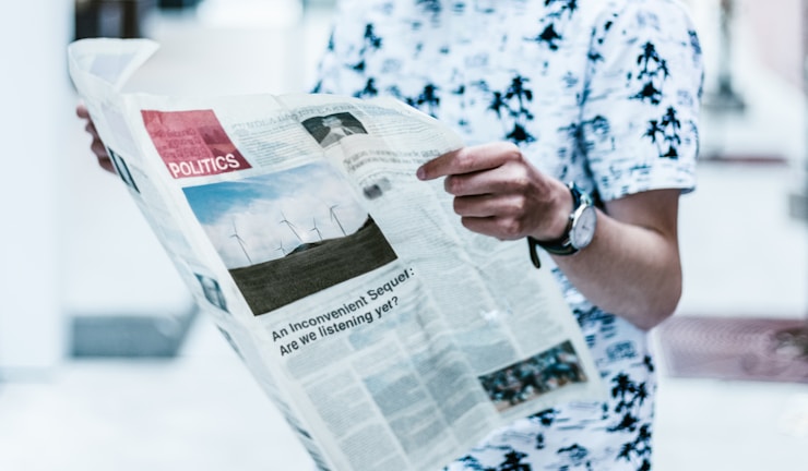 A person wearing a white shirt with black patterns is holding a newspaper. The newspaper is open and features a section labeled 'Politics,' along with a photo of wind turbines and a headline about climate change. The person is wearing a watch on their left wrist, and the background is slightly blurred.