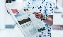 A person wearing a white shirt with black patterns is holding a newspaper. The newspaper is open and features a section labeled 'Politics,' along with a photo of wind turbines and a headline about climate change. The person is wearing a watch on their left wrist, and the background is slightly blurred.
