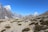 A group of hikers is trekking along a rocky and dry path surrounded by sparse vegetation in a mountainous region. Snow-capped peaks rise majestically in the background under a clear blue sky.