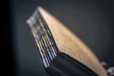 A close-up of a handmade book with visible stitching along the spine. The cover is textured with a metallic gold sheen, and the pages appear thick and slightly uneven, suggesting artisanal craftsmanship.