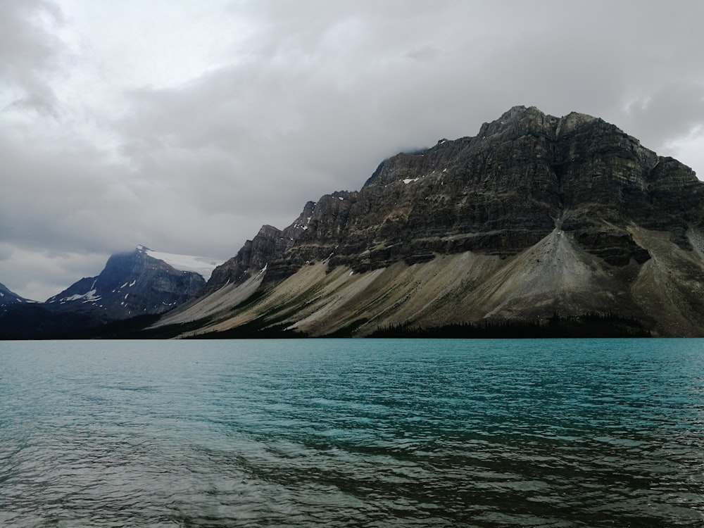 black and brown rocky mountain beside body of water under cloudy sky