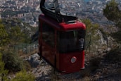A red cable car ascends above a rugged mountain landscape with a sprawling city in the background. Pine trees and rocky terrain are visible, with the urban area featuring dense clusters of buildings under a clear sky.