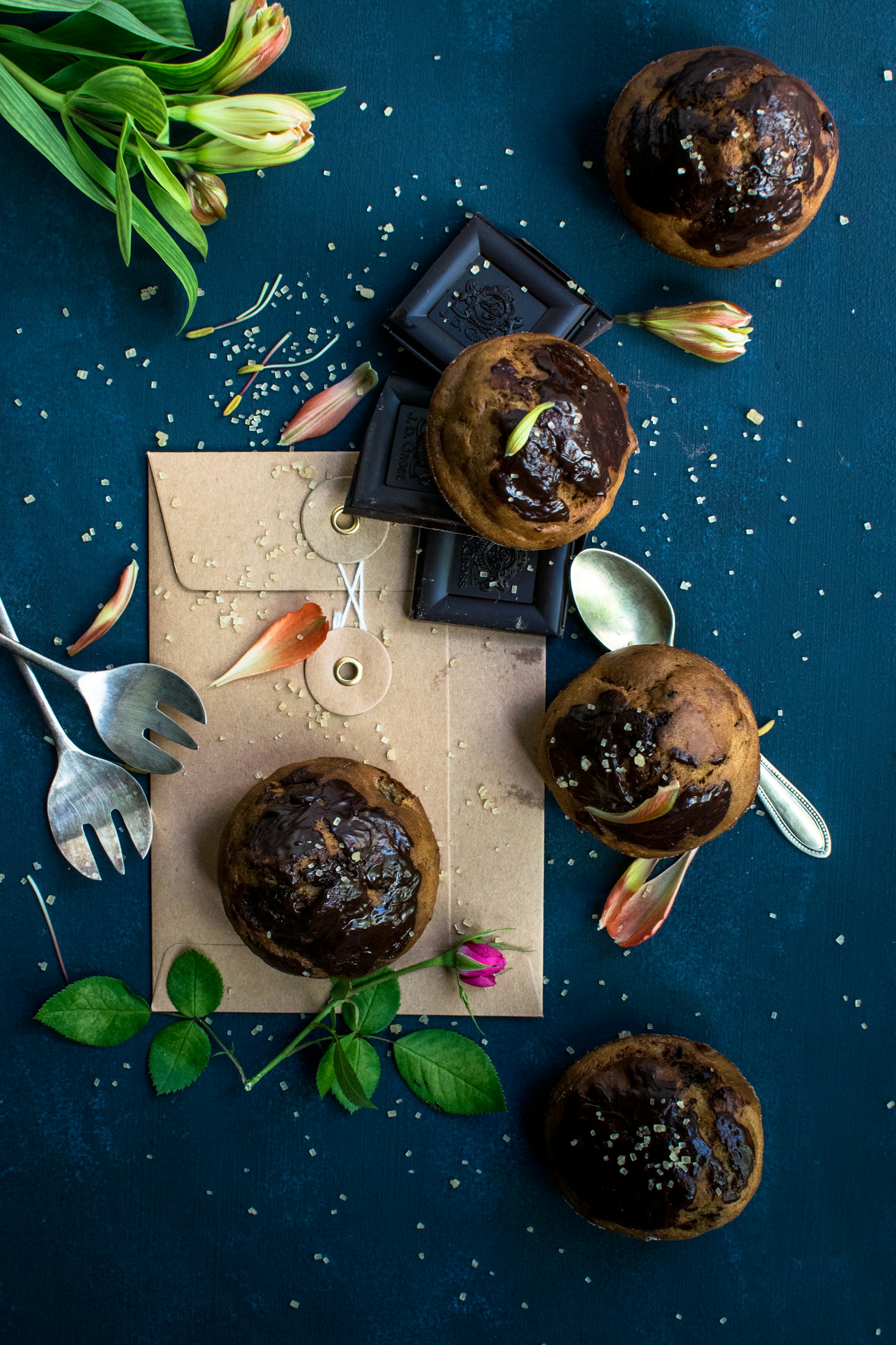 A beautifully arranged composition of baked chocolate muffins on a dark blue surface, surrounded by chocolate bars, tulip flowers, and an envelope. Scattered sugar granules and small pieces of petals accentuate the rustic and artistic display. Leafy green plants and metal cutlery add more elements to the scene.