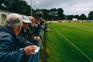 A group of people, mostly older men, is standing along the side of a football field, leaning against a metal fence. They appear to be engaged in conversation and watching a sporting event. The setting is outdoors, with lush green grass and trees in the background. The sky is overcast, and stadium lights are visible in the distance, suggesting an evening event.