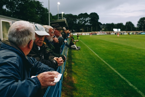 A group of people, mostly older men, is standing along the side of a football field, leaning against a metal fence. They appear to be engaged in conversation and watching a sporting event. The setting is outdoors, with lush green grass and trees in the background. The sky is overcast, and stadium lights are visible in the distance, suggesting an evening event.