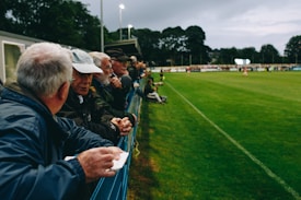 A group of people, mostly older men, is standing along the side of a football field, leaning against a metal fence. They appear to be engaged in conversation and watching a sporting event. The setting is outdoors, with lush green grass and trees in the background. The sky is overcast, and stadium lights are visible in the distance, suggesting an evening event.