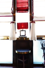 A stylish interior with a round stool at the center, featuring a dark cushion and golden trim. Behind the stool is a lamp with a pleated red shade, positioned on a narrow table situated between large windows. The windows are dressed with red and white curtains that allow soft, diffused light into the space.