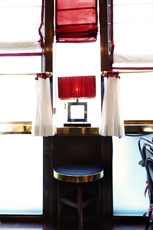 A stylish interior with a round stool at the center, featuring a dark cushion and golden trim. Behind the stool is a lamp with a pleated red shade, positioned on a narrow table situated between large windows. The windows are dressed with red and white curtains that allow soft, diffused light into the space.