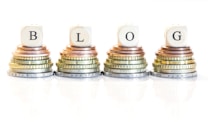 Stacks of coins arranged in four piles, each topped with a wooden block bearing a letter, collectively spelling 'BLOG'. The coins vary in size and color, ranging from copper to silver and gold.