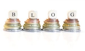 Stacks of coins arranged in four piles, each topped with a wooden block bearing a letter, collectively spelling 'BLOG'. The coins vary in size and color, ranging from copper to silver and gold.