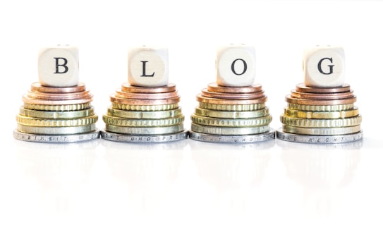 Stacks of coins arranged in four piles, each topped with a wooden block bearing a letter, collectively spelling 'BLOG'. The coins vary in size and color, ranging from copper to silver and gold.