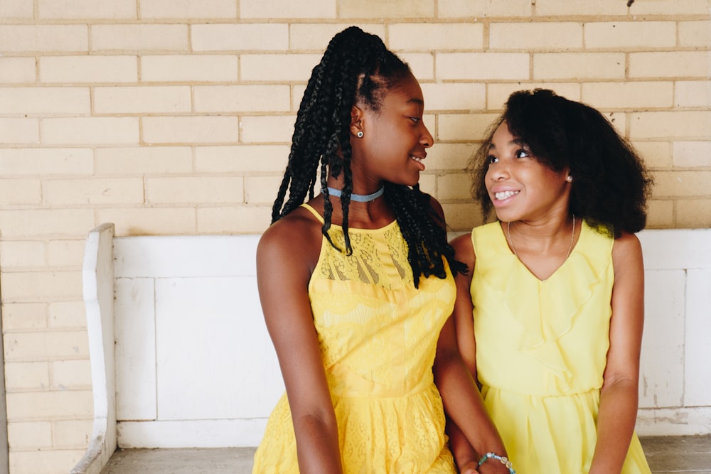 two girl's in yellow sleeveless dresses sitting on white wooden bench during daytime