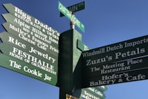 A green multi-directional signpost against a clear blue sky, displaying various local business names and street names. The names include restaurants, jewelry stores, and bakeries. The businesses are advertised with traditional fonts and are possibly in an area with a European or Bavarian theme.