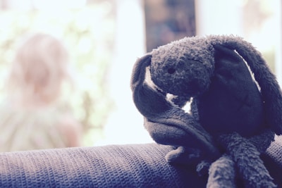 A plush toy, possibly a rabbit, with long ears and a soft, textured appearance sits on a dark textured surface. In the background, there is an out-of-focus view of a child with blonde hair, adding a sense of depth to the image.