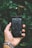 A hand holding a smartphone with the screen displaying camera settings. The phone screen appears cracked. The wrist of the hand is adorned with a silver watch. The background consists of lush green leaves, creating a natural setting.