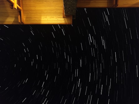 Long exposure photograph capturing star trails in the night sky with a section of a wooden structure visible, possibly a deck or rooftop, in the upper frame.