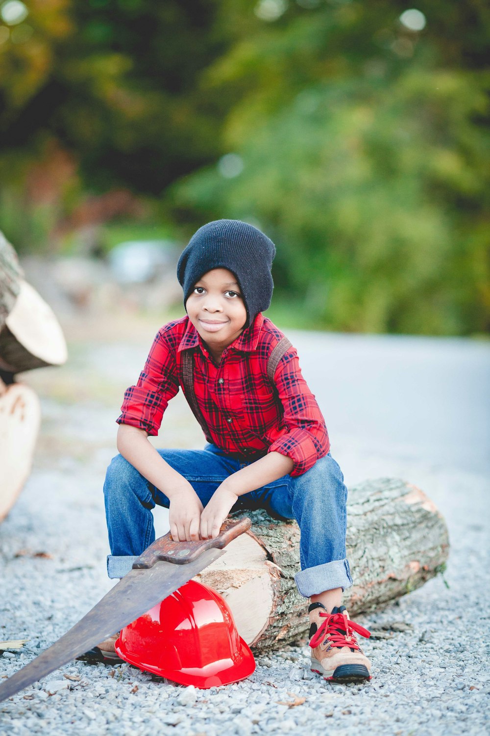 丸太の上に座り、赤いヘルメットの上に手のこぎりを持っている少年