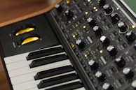 A close-up of an electronic music synthesizer, featuring a keyboard with black and white keys and a control panel filled with various knobs, buttons, and switches. The knobs are metallic and there are yellow LED lights indicating the synthesizer's operation.