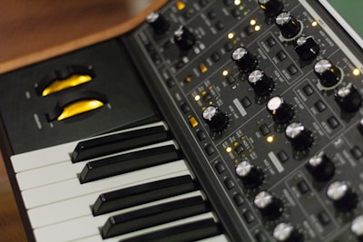 A close-up of an electronic music synthesizer, featuring a keyboard with black and white keys and a control panel filled with various knobs, buttons, and switches. The knobs are metallic and there are yellow LED lights indicating the synthesizer's operation.