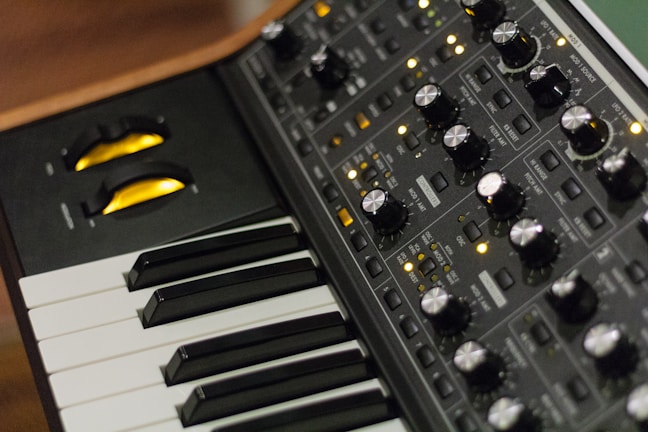 A close-up of an electronic music synthesizer, featuring a keyboard with black and white keys and a control panel filled with various knobs, buttons, and switches. The knobs are metallic and there are yellow LED lights indicating the synthesizer's operation.