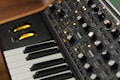 A close-up of an electronic music synthesizer, featuring a keyboard with black and white keys and a control panel filled with various knobs, buttons, and switches. The knobs are metallic and there are yellow LED lights indicating the synthesizer's operation.