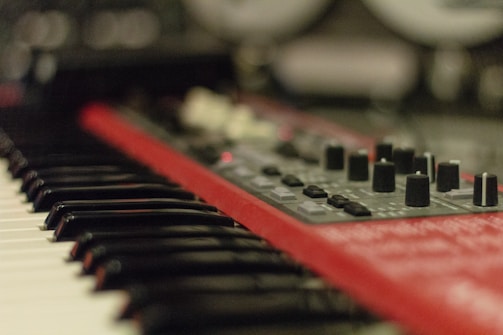 A close-up of a red synthesizer with black and white piano keys visible in the foreground. Several knobs and buttons are present on the synthesizer's control panel with a soft focus effect creating a blurred background.