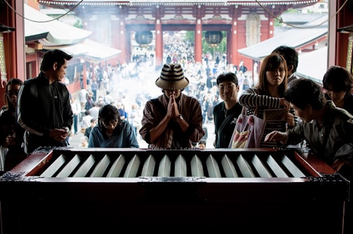 A group of people are gathered around a large offering box inside a temple. In the middle, one person in a striped hat is praying with hands clasped together. Other individuals around are engaged in various activities, such as reading and talking. In the background, there is a large crowd visible through the temple entrance, surrounded by traditional architecture and smoke rising from incense.