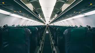 An empty airplane cabin featuring rows of seats with overhead compartments. The perspective shows the narrow aisle leading towards the far end of the aircraft. The lighting is subdued, with small illuminated signs visible above the seats.