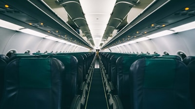An empty airplane cabin featuring rows of seats with overhead compartments. The perspective shows the narrow aisle leading towards the far end of the aircraft. The lighting is subdued, with small illuminated signs visible above the seats.