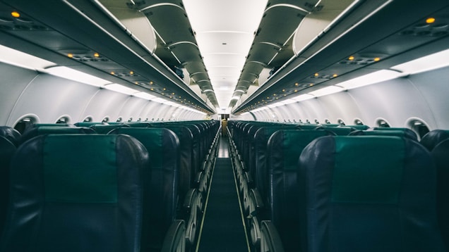 An empty airplane cabin featuring rows of seats with overhead compartments. The perspective shows the narrow aisle leading towards the far end of the aircraft. The lighting is subdued, with small illuminated signs visible above the seats.