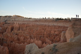 A dramatic landscape featuring a vast canyon with intricate rock formations. The scene is bathed in soft daylight, highlighting the earthy tones of the terrain. Two people stand on the edge of a cliff, suggesting a moment of exploration and adventure. The horizon features a clear sky with scattered clouds.