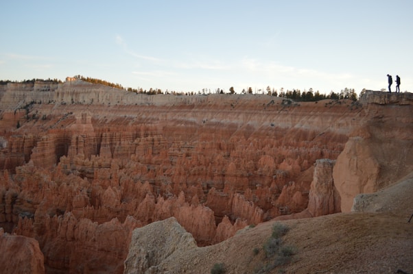 A dramatic landscape featuring a vast canyon with intricate rock formations. The scene is bathed in soft daylight, highlighting the earthy tones of the terrain. Two people stand on the edge of a cliff, suggesting a moment of exploration and adventure. The horizon features a clear sky with scattered clouds.