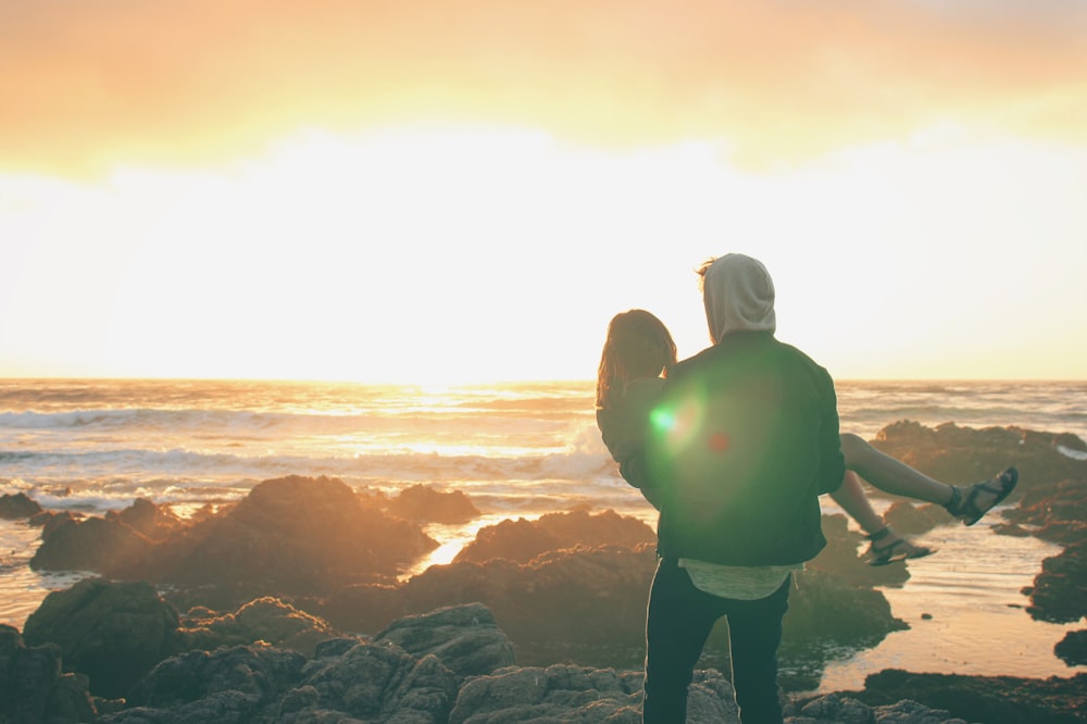 man carrying the woman on top of cliff near ocean during sunset