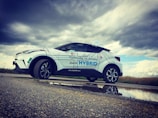 A white hybrid SUV is parked on a rural road next to a puddle, reflecting its image beneath a cloudy sky. The car has promotional text in a foreign language, with large letters highlighting the word 'HYBRID.' The landscape in the background is grassy with overcast weather conditions.