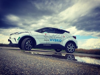 A white hybrid SUV is parked on a rural road next to a puddle, reflecting its image beneath a cloudy sky. The car has promotional text in a foreign language, with large letters highlighting the word 'HYBRID.' The landscape in the background is grassy with overcast weather conditions.