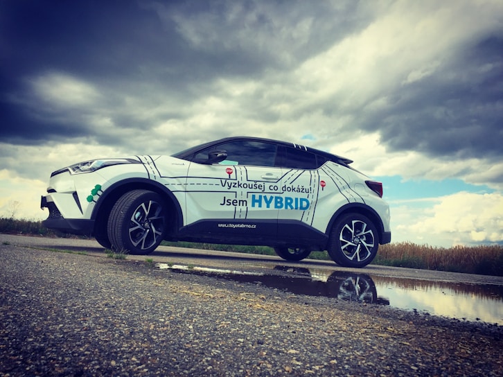 A white hybrid SUV is parked on a rural road next to a puddle, reflecting its image beneath a cloudy sky. The car has promotional text in a foreign language, with large letters highlighting the word 'HYBRID.' The landscape in the background is grassy with overcast weather conditions.