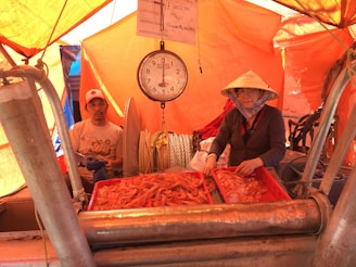 Two individuals are sorting a pile of fresh shrimps into red trays aboard a fishing vessel. The setting is under an orange tarp, suggesting a market or harbor environment. A mechanical dial scale is hanging above the trays, indicating the weight of the catch. The person on the right is wearing a traditional Asian conical hat, adding a cultural aspect to the scene.