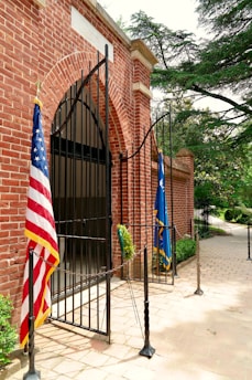 An open wrought iron gate set in a red brick archway leads into a shaded area. Two flags, including an American flag, are displayed prominently at the entrance. The scene is surrounded by greenery, with trees visible in the background.