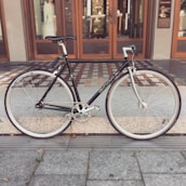 A black bicycle with drop handlebars is parked on a tiled walkway in front of a wooden-framed glass door. The bike has white tires and a clean, sleek design.