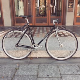 A black bicycle with drop handlebars is parked on a tiled walkway in front of a wooden-framed glass door. The bike has white tires and a clean, sleek design.