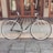 A black bicycle with drop handlebars is parked on a tiled walkway in front of a wooden-framed glass door. The bike has white tires and a clean, sleek design.