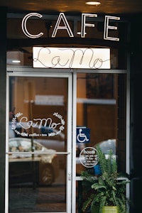 A cafe entrance with a glass door displaying the name La Mo, surrounded by decorative leaves, and offering coffee and tea. Above the door is a large illuminated sign spelling CAFE. A blue wheelchair accessibility sign is visible next to the door. A potted plant sits in front of the entrance. Business hours are posted on the door.